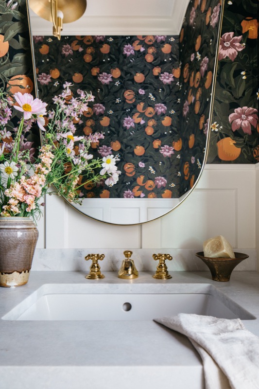 Bathtub beneath a round brass mirror and moody floral wallpaper · Oak Park Main Level Renovation by Leah Phillips Interiors